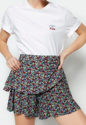Frau trägt ein weißes T-Shirt mit hochgekrempelten Ärmeln und dem Text "Paris 1987" sowie einen bunten, mehrlagigen Blumenrock, Hände in die Hüften gestützt.