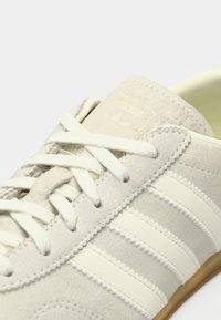 Jasnoszary zamszowy sneaker z kremowymi paskami, teksturowanymi akcentami i płaskimi sznurowadłami. Na języku znajduje się naszywka z logo w stylu vintage.