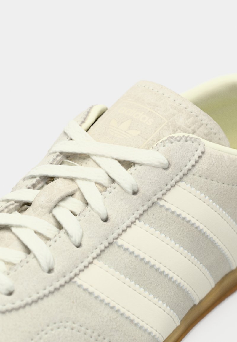 Light gray suede sneaker with cream stripes, textured accents, and flat laces. Features a vintage-style logo patch on the tongue.