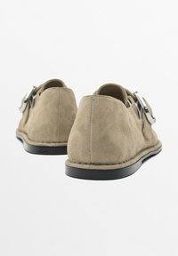 Suede slip-on shoes in a muted beige tone, featuring a round toe and a silver metal buckle accent on the side. Black outsole adds contrast.