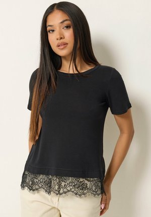 Jonge vrouw met lang steil haar, die een zwarte top met korte mouwen draagt met een kanten zoom, gecombineerd met lichtgekleurde broek.