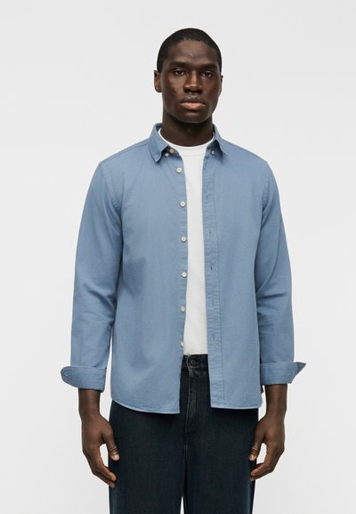 Chemise bleu clair en coton à boutons, avec col, manches retroussées et un t-shirt blanc en dessous, associée à un jean en denim foncé.
