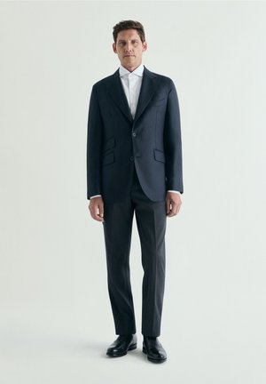 Hombre de pie con un traje azul marino oscuro, camisa blanca y zapatos de vestir negros contra un fondo claro y liso.