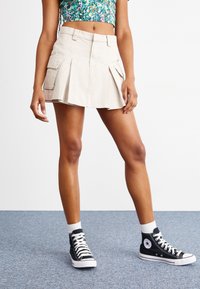 Beige, rakott, mini szoknya, két oldalsó zsebből készült, pamutból. Fekete, magas szárú sportcipővel és fehér bokazoknival párosítva.