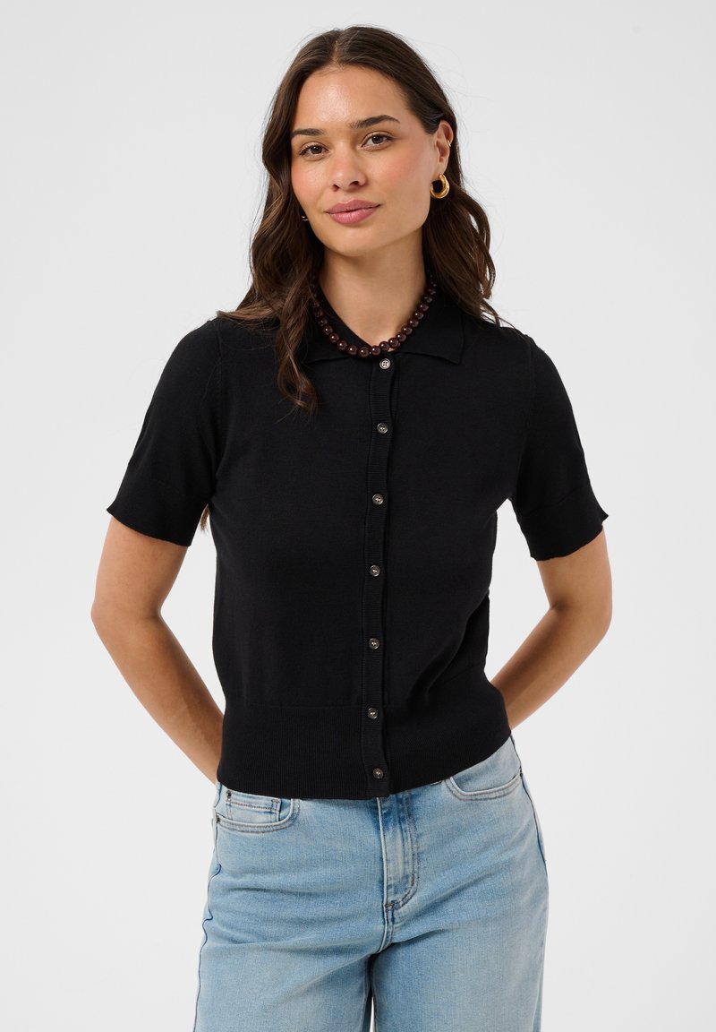Femme aux longs cheveux bruns portant un haut noir à manches courtes boutonné, un jean bleu, des boucles d’oreilles créoles dorées et un collier foncé à perles.