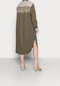 Robe verte olive avec un col à motifs plaid et des broderies détaillées dans le dos. Présente des fentes latérales et une coupe longue et ample.