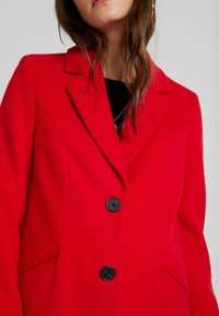 Frau in einem roten, geknöpften Mantel mit schwarzen Knöpfen, teilweise sichtbare Halskette und lockigem, braunem Haar vor einem hellen Hintergrund.