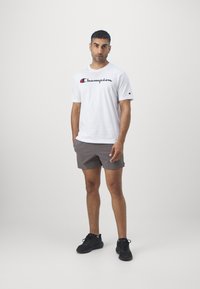 Vit bomull T-shirt med en svart och röd "Champion"-logotyp, i kombination med grå shorts och svarta sportskor. Slät struktur, avslappnad design.