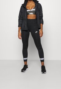 Czarna sportowa kurtka, metalicznie szary stanik sportowy, czarne legginsy z subtelnym wzorem i logo. Czarne sneakersy z białymi akcentami na podeszwie.