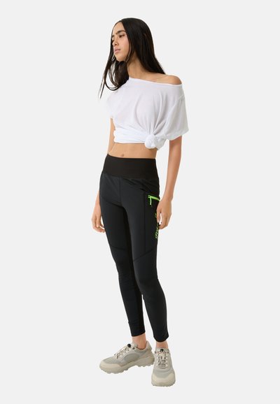 Schwarze Sportleggings mit einer grünen Reißverschlusstasche, kombiniert mit einem weißen, geknoteten, kurz geschnittenen T-Shirt und hellgrauen Sneakers.