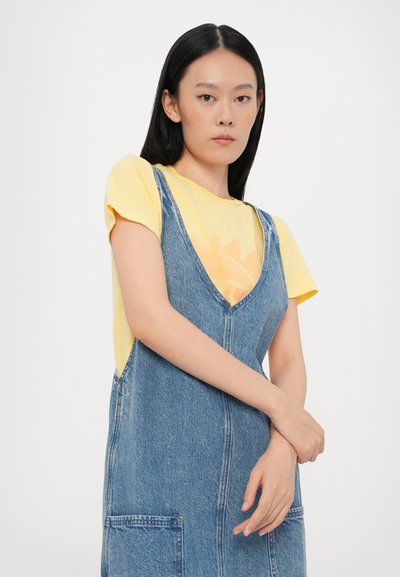 Robe salopette en denim avec un décolleté en V plongeant, de couleur bleue, dotée de deux poches avant, portée par-dessus un T-shirt graphique jaune.
