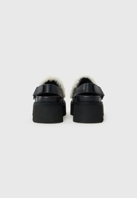 Zwarte platform sandalen met witte fuzzy voering, verstelbare klittenbandriemen en dikke zolen, bekeken van achteren op een witte achtergrond.