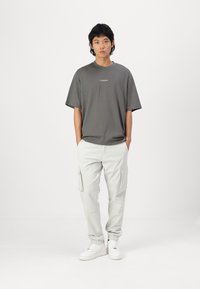 Šedé oversized tričko s krátkým rukávem a logem. Světle šedé cargo kalhoty s několika kapsami. Bílé tenisky dotvářejí celý outfit.