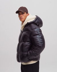 Joven con una chaqueta negra acolchada brillante con capucha de forro polar blanca y una gorra marrón, de pie de lado sobre un fondo liso.