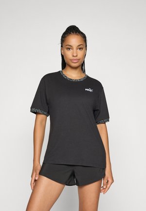 Kvinne iført en svart Puma t-skjorte med mønstrede detaljer og matchende sorte shorts, stående mot en enkel lys bakgrunn.