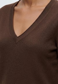 Gros plan d’une personne portant un pull marron à col en V montrant l’encolure et la partie supérieure de la poitrine.