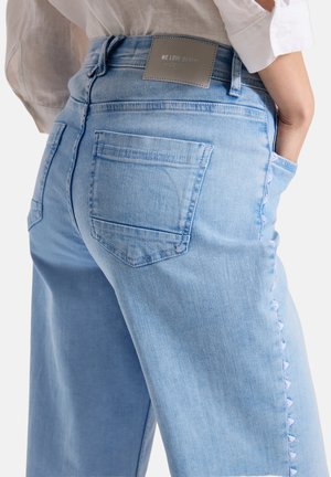 Lichtblauwe spijkerbroek met achterzak en zijknopjes, gedragen met een wit shirt met lange mouwen, ingestopt.