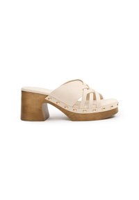 Chanclas beige con diseño de tiras cruzadas, grueso tacón cuadrado de tono dorado y acentos de tachuelas decorativas a lo largo de la suela. Acabado de material texturizado.