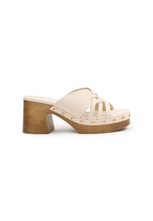 Chanclas beige con diseño de tiras cruzadas, grueso tacón cuadrado de tono dorado y acentos de tachuelas decorativas a lo largo de la suela. Acabado de material texturizado.