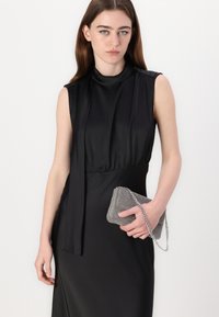 Black sleeveless dress with a high collar and draped accents, paired with a metallic mesh clutch featuring a chain strap.