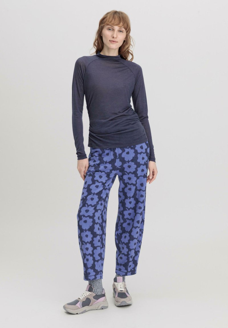 Langärmliges, dunkelblaues Oberteil mit einer taillierten Passform, kombiniert mit lockeren, floral gemusterten blauen Hosen. Graue Sneaker mit rosa Akzenten runden das Outfit ab.