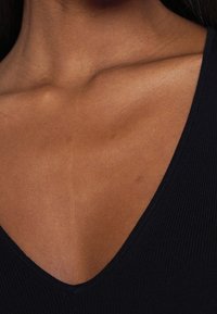 Schwarzer gerippter Stoff mit einem tiefen V-Ausschnitt, der glatte Haut am Schlüsselbein und den Schultern zeigt. Subtle Struktur, minimalistische Details.