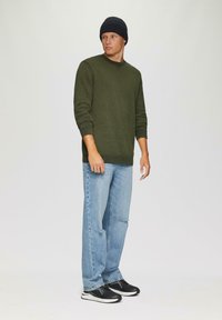 Groene gebreide trui met ronde hals, gecombineerd met lichtblauwe loszittende jeans en zwarte sneakers. Het model draagt een zwarte beanie.