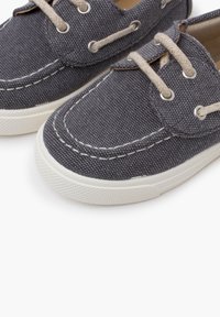 Dunkelgraue Segelschuhe aus Canvas mit beigen Schnürsenkeln, silbernen Ösen und weißen Gummisohlen. Sie weisen sichtbare Nähte und eine strukturierte Oberfläche auf.