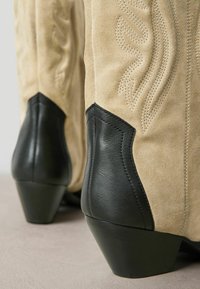 Tan suède cowboylaarzen met een zwarte leren hak, voorzien van ingewikkelde stiksels en een kegelvormig hakontwerp. Gelaagde afwerking.