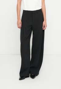 Pantalon noir à jambes larges avec plis, présentant un devant plat, des passants pour ceinture et un tissu léger et lisse. Porté avec un haut blanc et des escarpins noirs.