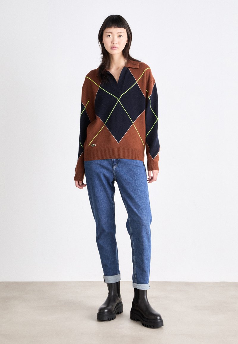 Pull en tricot à motifs argyle brun et marine avec des accents verts, un col, et un ourlet côtelé. Porté avec un jean bleu retroussé et des bottes noires chunky.