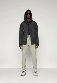 Črna kapuco jakna z zadrgo spredaj in stranskimi žepi, v kombinaciji s svetlo zelenimi hlačami. Model nosi sončna očala in superge, stoji na enobarvni podlagi.