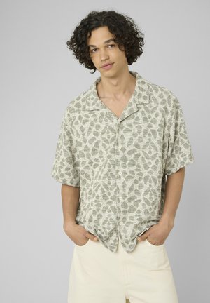 Jeune homme aux cheveux bouclés portant une chemise à manches courtes à motifs et un pantalon clair, debout les mains dans les poches.