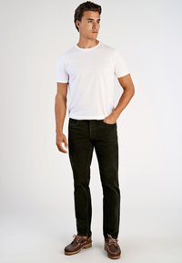 Camiseta de algodón blanca, pantalones de pana negra, zapatos marrones con cordones. Modelo de pie recto, mano en el bolsillo, fondo neutral.