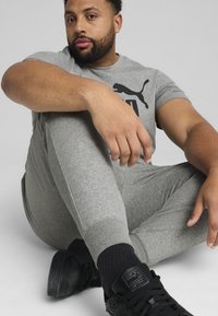 Grå atletisk sæt med en tætsiddende t-shirt med et sort logo og matchende joggers. Sorte sneakers fuldender looket på en grå baggrund.