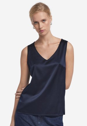 Kvinde med lyst hår iført sølvfarvede creoler og en ærmeløs, marineblå satin top med V-hals samt mørkeblå jeans.