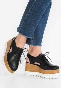 Chaussures à plateforme en cuir noir avec découpes, design à lacets, semelle intermédiaire beige et semelle blanche crantée. Portées avec un jean en denim bleu effiloché.