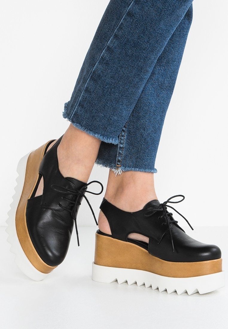 Chaussures à plateforme en cuir noir avec découpes, design à lacets, semelle intermédiaire beige et semelle blanche crantée. Portées avec un jean en denim bleu effiloché.