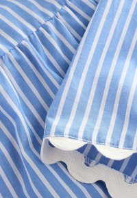 Blåt og hvidt stribet stof med en tekstureret bølgekant. Materialet virker blødt og let. Mønstrene er jævnt fordelt.