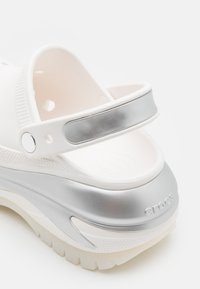 Balti Crocs sandalai su sidabro akcento dirželiu, tekstūruota pakyla ir tvirtu padu su sukibimo raštais. Lygus gumos paviršius.