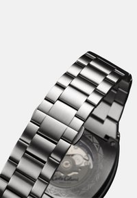 Silver metal watch band with flat links, brushed finish, and a clasp. The back features a transparent case showcasing the inner mechanics.