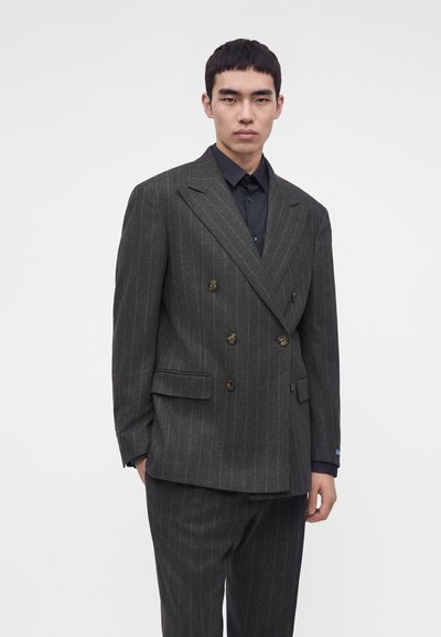 Homme portant un costume croisé gris foncé à fines rayures avec une chemise noire, debout devant un fond clair uni.