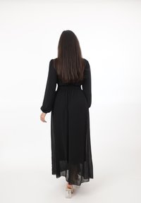 Kobieta o długich włosach, ubrana w długą czarną suknię i przezroczyste sandały na obcasie, stoi plecami do kamery na białym tle.