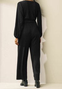 Jumpsuit noir plissé à manches longues, taille cintrée et bas à jambe large ; texture lisse ; porté avec des bottes à talons noires.