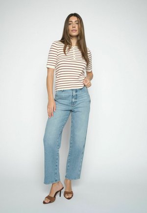 Frau mit langen braunen Haaren, die ein kurzärmeliges weiß-rotes gestreiftes Shirt, hellblaue hoch taillierte Jeans und braune Sandalen mit Absatz trägt.