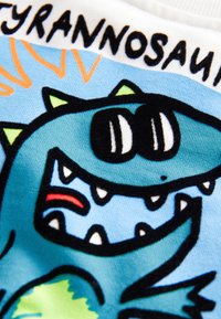 Tricou cu un dinozaur desen animat, albastru cu contur negru, dinți ascuțiți și țepi verzi, pe un fundal albastru deschis.