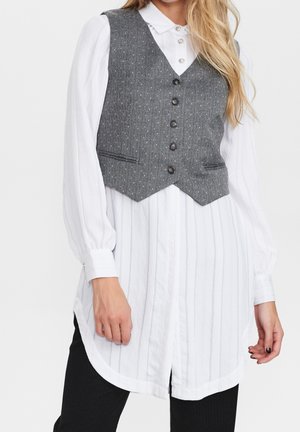 Femme portant un gilet gris boutonné sur une longue blouse blanche texturée à manches bouffantes et un pantalon noir, devant un fond uni.