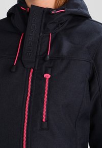 Granatowa kurtka z kapturem, wykonana z teksturowanej tkaniny, z różowymi zamkami błyskawicznymi i naszywką z logo z przodu. Charakterystyczne akcenty sznureczków.