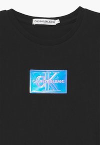Czarna bawełniana koszulka z błyszczącą holograficzną prostokątną naszywką z wytłoczonymi srebrnymi literami „CK” i napisem „CALVIN KLEIN JEANS”. Okrągły dekolt.