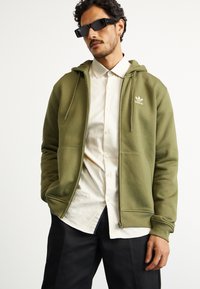 Manta com fecho éclair na cor verde oliva, com um acabamento texturado, bolsos frontais e logótipo branco. Combinada com uma camisa de botão, óculos pretos e calças escuras.